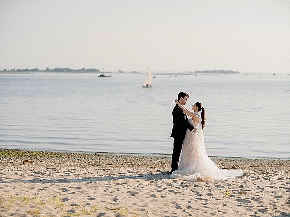 Couple portrait of bride in strapless wedding dress and veil embracing groom in tuxedo on a sandy beach with ocean and sailboat behind