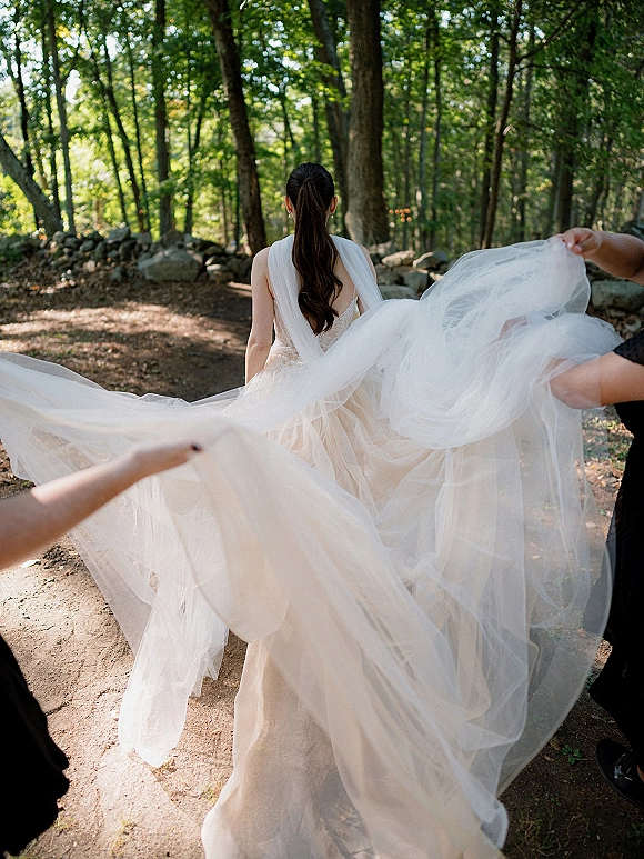 Bride portrait from behind in a tulle wedding dress train and long veil, walking a woodland path beside a stone wall