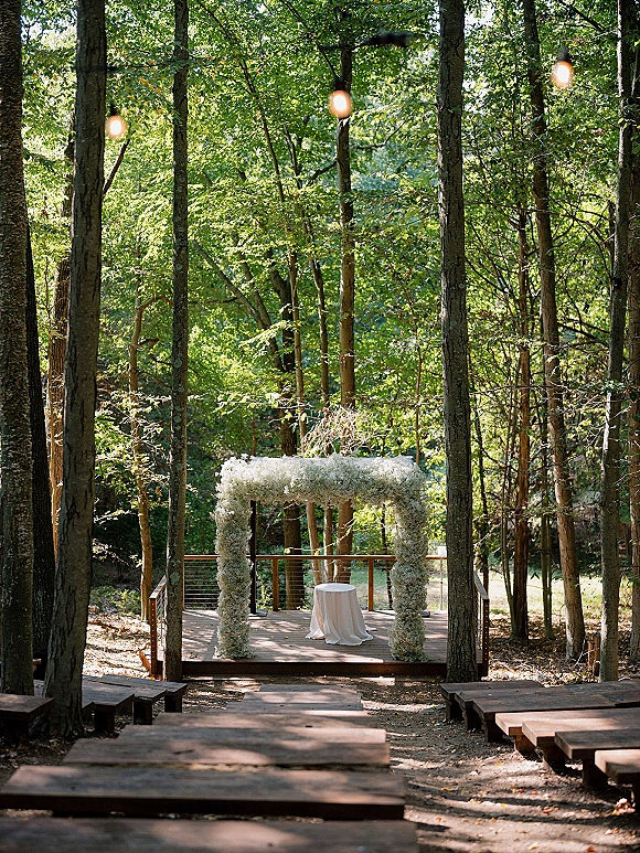 Ceremony setup with a baby’s breath floral arch and string lights, wooden benches lining a dirt path in a forest clearing