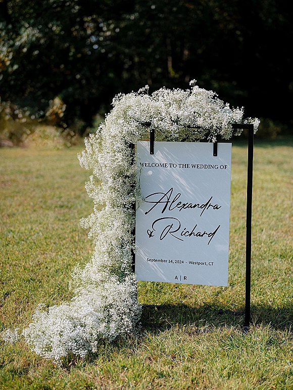 Wedding welcome sign framed with baby’s breath flowers on a black metal stand, set on a grass lawn with trees behind