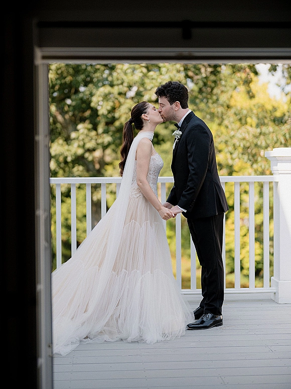 Wedding kiss portrait of bride and groom kissing on a wooden porch, her cathedral veil flowing over a tulle skirt beside a white railing