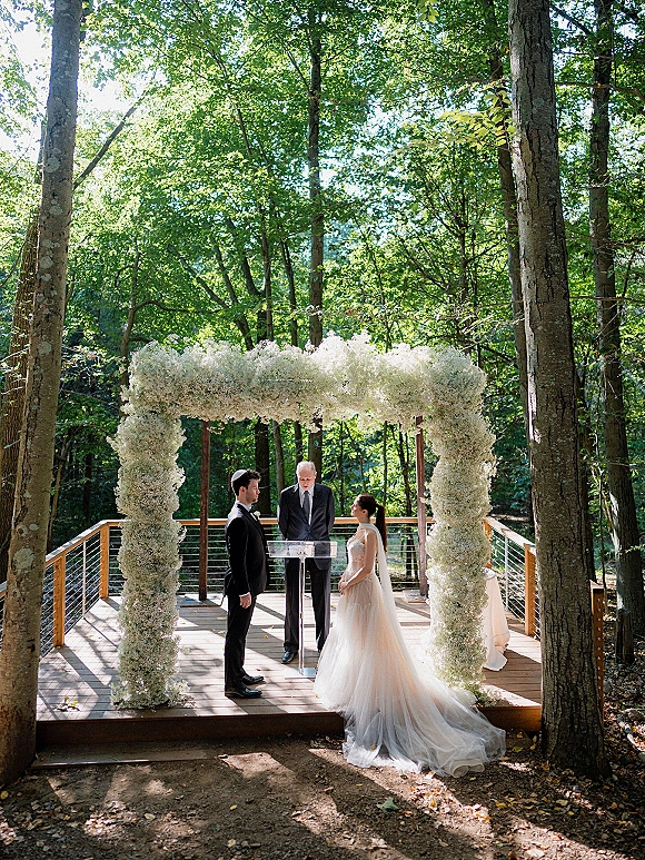 Wedding ceremony under a floral arch with bride in long veil and train facing groom in tuxedo on a sunlit wooden deck in forest