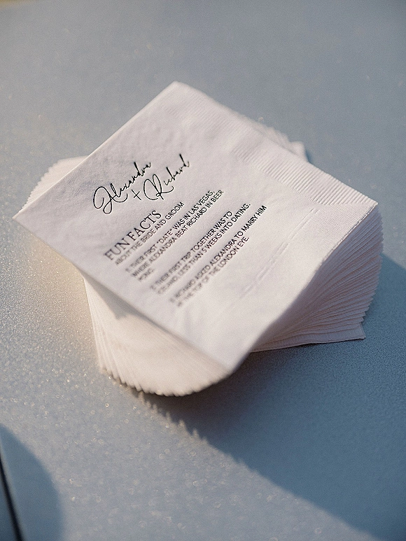 Wedding cocktail napkins with personalized wedding napkins script lettering stacked neatly on a light blue tabletop, showing custom printed text