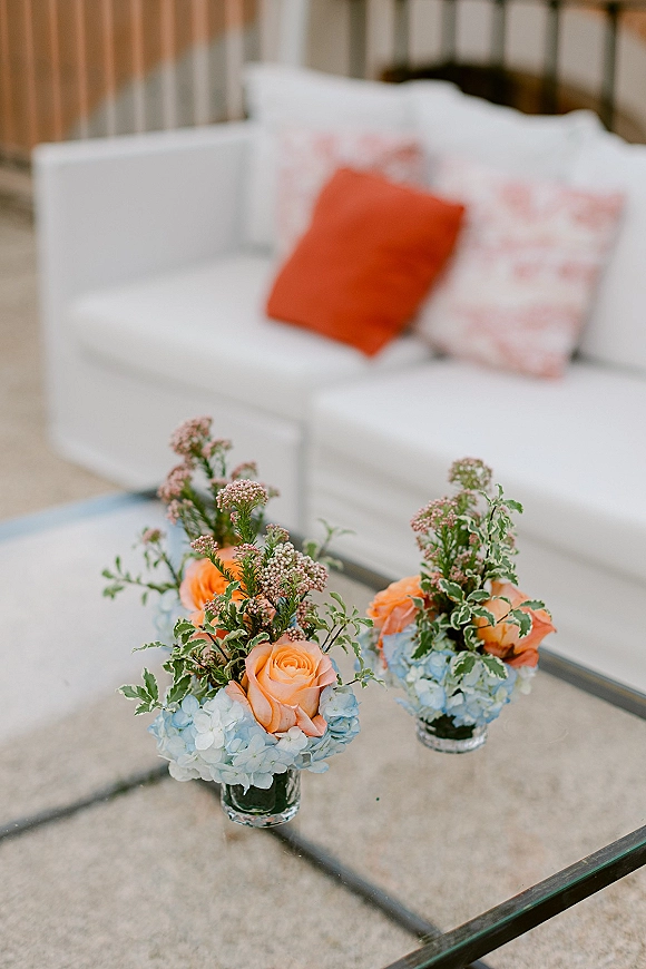 Wedding floral centerpieces of orange and blue wedding flowers in small glass vases on a glass coffee table beside a white sofa in a lounge area