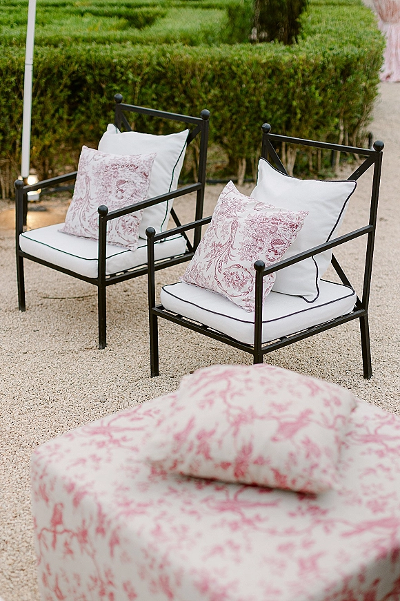 Wedding lounge seating with black metal chairs and white cushions, patterned throw pillows, and an upholstered ottoman on a gravel patio by hedges