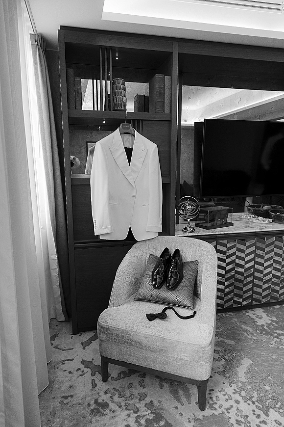 Groom attire details with a white tuxedo jacket on a suit hanger, black patent dress shoes and bow tie arranged in a hotel suite room