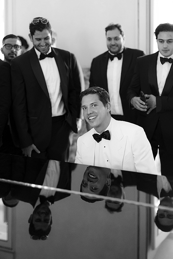 Groom portrait in a white tuxedo jacket and bow tie, seated at a piano with groomsmen in tuxedos and sunglasses in an indoor room