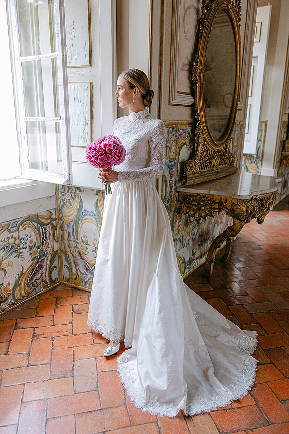 Bridal portrait of a bride by window holding a pink bouquet in a high-neck lace gown with drop earrings, framed by an ornate gold mirror.