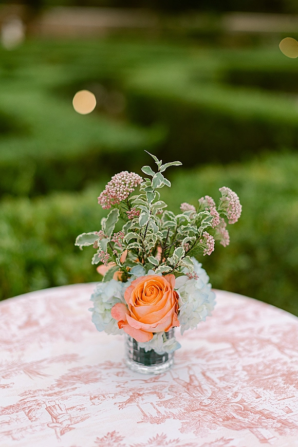 Wedding centerpiece with an orange rose and pale blue hydrangea in a glass vase on a pink toile cocktail table, garden hedges behind