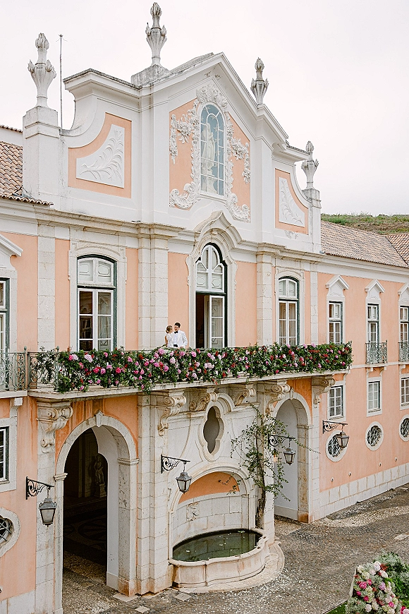 Couple portrait of bride and groom on a wedding balcony portrait, embracing by a pink floral garland on an ornate historic facade with arches