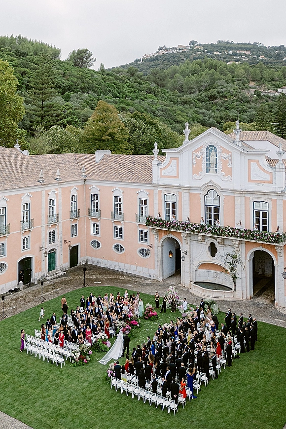 Outdoor wedding ceremony with white chairs circling an aisle runner and floral arch in a historic courtyard with arched colonnade backdrop