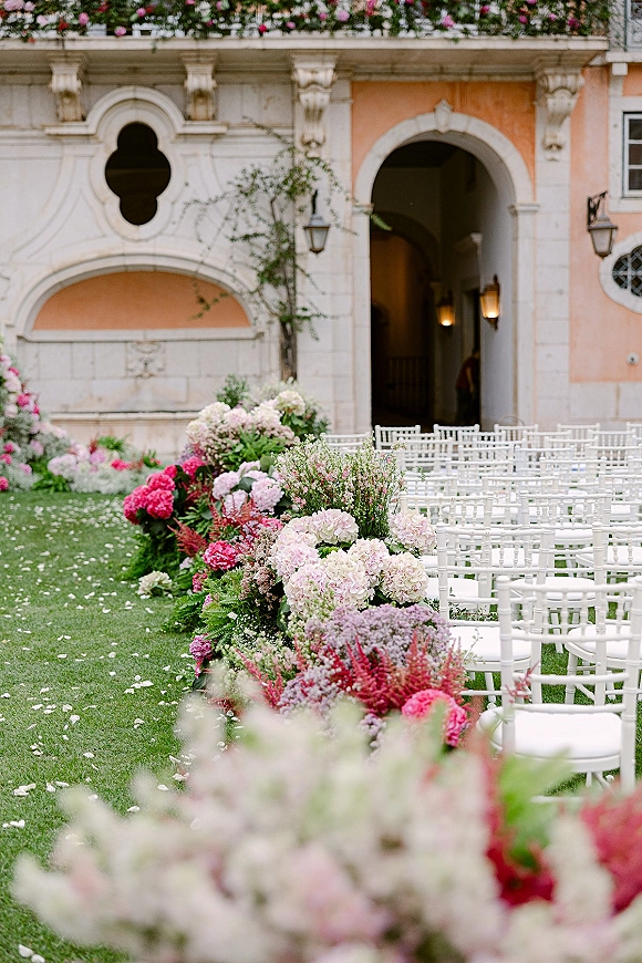 Ceremony aisle decor with wedding aisle flowers, hydrangeas and pink blooms lining white chairs with petals in a courtyard by an arched doorway