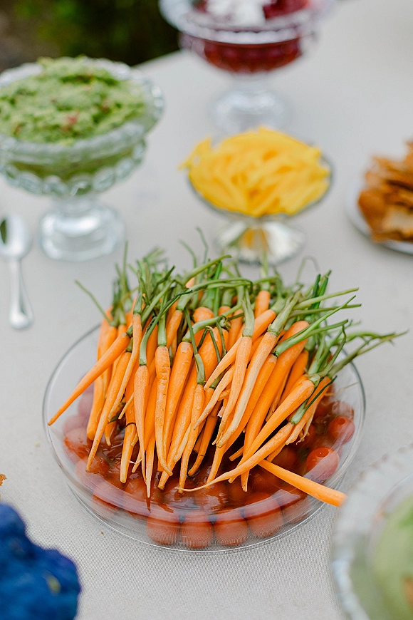 Wedding appetizer table with a crudite platter of baby carrots and dips in glass bowls, set on a tablecloth with greenery behind