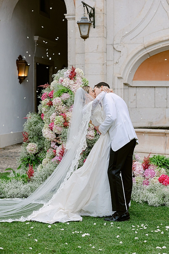 Wedding kiss as bride in lace dress and cathedral veil kisses groom in white jacket before stone archway with hydrangea florals and petals