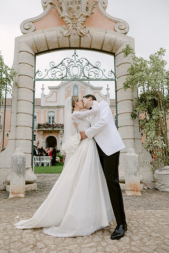 Wedding kiss portrait of the bride and groom kissing under an ornate stone archway, her veil flowing beside his white tuxedo jacket.
