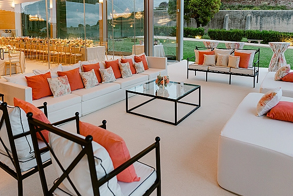 Wedding lounge area with white sofas and orange throw pillows around a glass coffee table, set by glass walls overlooking a garden patio lawn