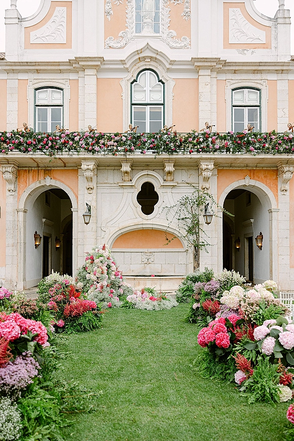 Ceremony aisle decor with floral lined wedding aisle of hydrangeas and roses bordering white chairs in a palace courtyard with arched doors