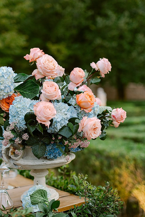 Wedding centerpiece with peach roses and blue hydrangeas in a white compote, set on a wooden table with glassware on a garden lawn