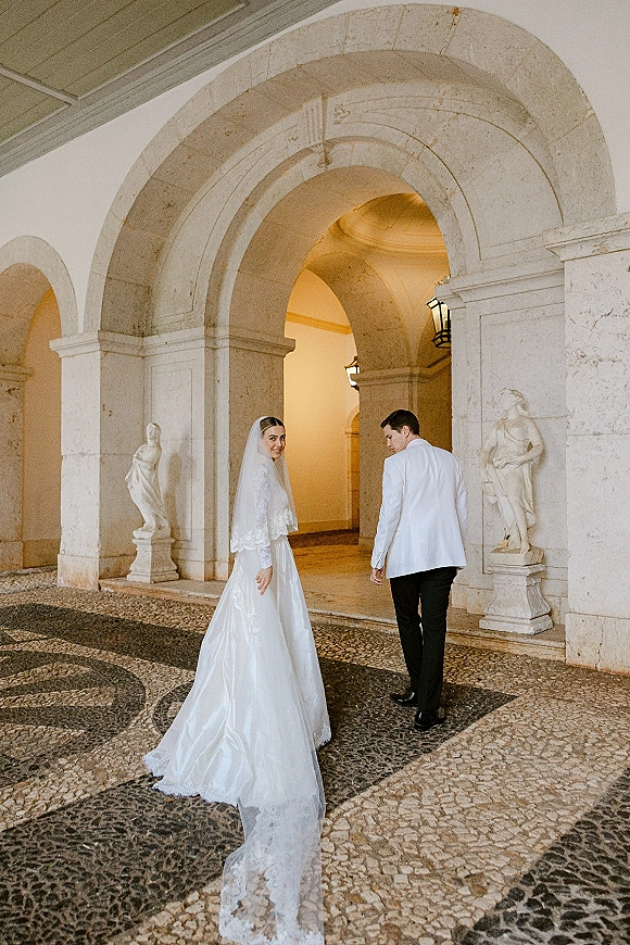 Wedding couple portrait with bride looking back as she and groom walk under stone archways, her cathedral veil trailing by statues and lanterns