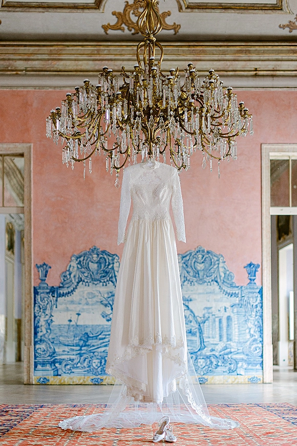 Wedding dress with long sleeve wedding dress lace bodice hanging from a crystal chandelier, veil draped over tulle skirt in ornate room
