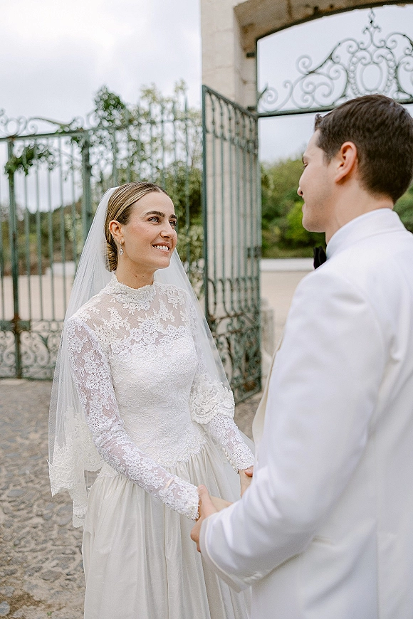 First look moment as bride and groom hold hands, she smiles in lace dress and cathedral veil by wrought iron gates on cobblestones