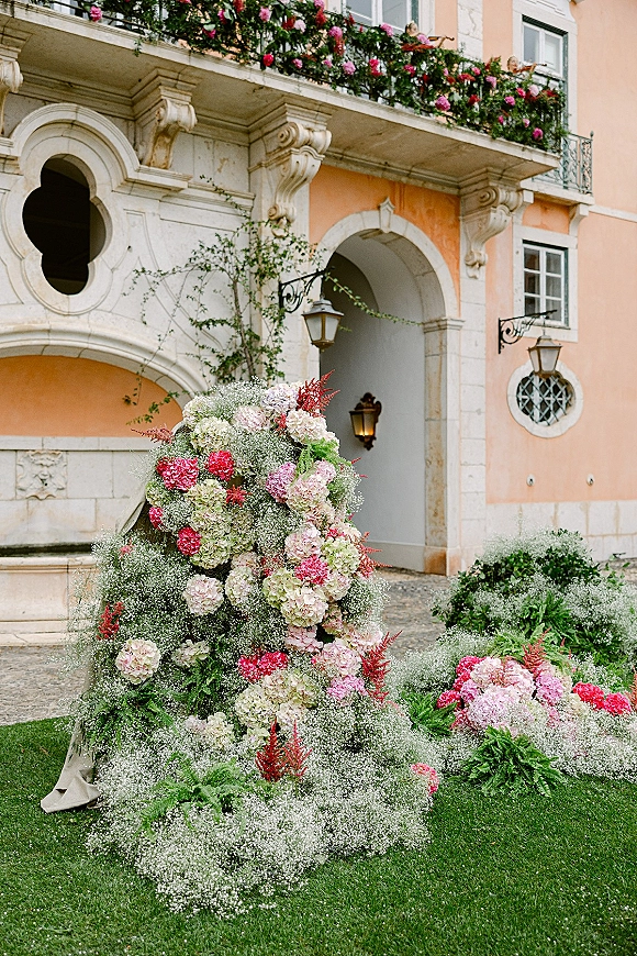 Wedding ceremony flowers with ceremony floral installation of hydrangeas and baby’s breath, with draped fabric by an arched stone doorway