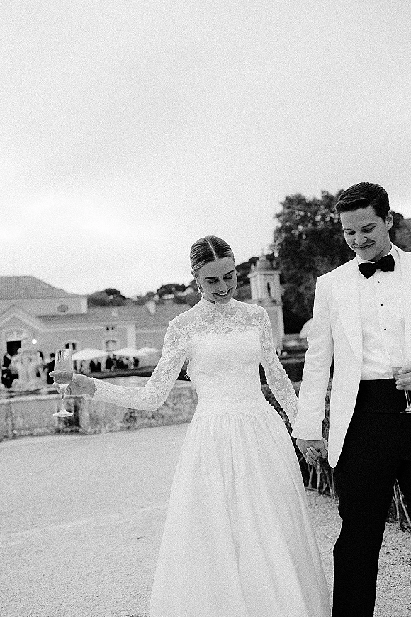 Couple portrait of newlyweds holding hands, bride in high-neck lace gown beside groom in white tux on an estate terrace by stone wall