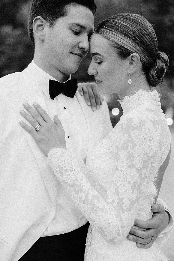 Wedding couple portrait in a black and white wedding photo, close up as they touch foreheads, bride’s ringed hand on groom in white tuxedo by bokeh greenery
