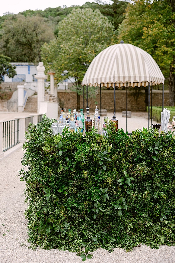 Wedding bar setup with an outdoor wedding bar wrapped in greenery, liquor bottles and drink dispensers under a striped canopy in a gravel courtyard terrace