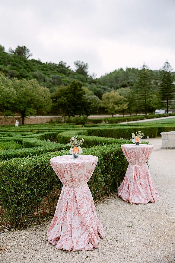 Cocktail table decor with pink patterned tablecloths and floral centerpieces on highboy tables along a gravel garden path under cloudy skies