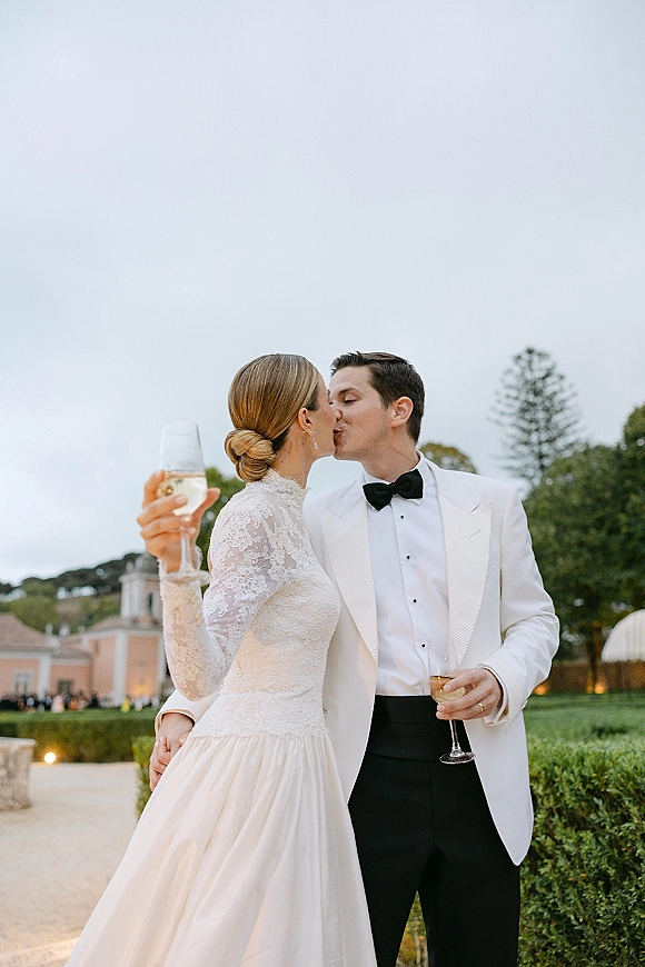 Wedding kiss portrait of bride and groom toasting with champagne, her long-sleeve lace dress and his white dinner jacket in an estate garden walkway