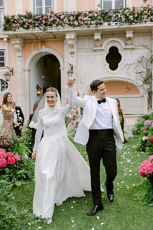 Wedding recessional as bride and groom hold hands walking a rose petal aisle on a garden lawn, guests cheering by a historic facade with archway