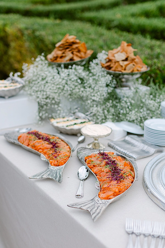 Wedding cocktail hour wedding appetizer display with salmon platters on silver fish trays, baby’s breath accents, and plates on a white linen table on a green lawn