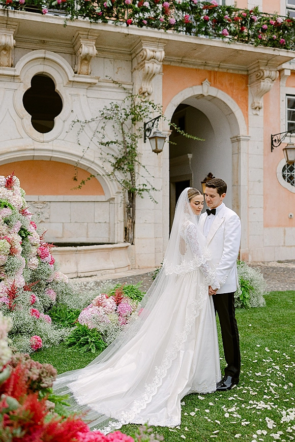 Couple portrait of bride and groom holding hands, her cathedral veil and lace train on grass by a villa arch with hydrangeas
