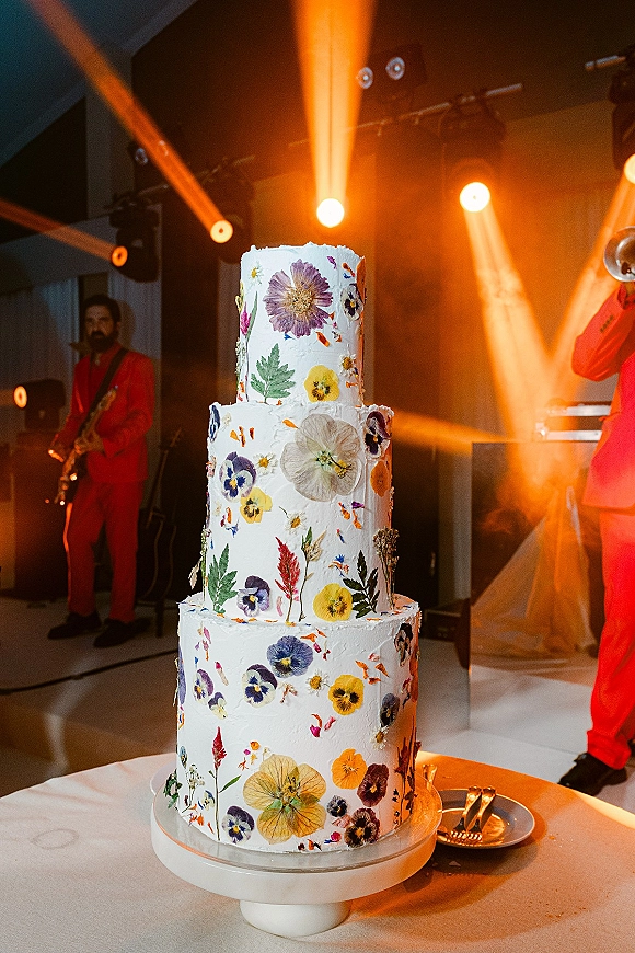 Wedding cake with pressed flower wedding cake design on a cake stand, with knife and server set under stage lighting near the band on the dance floor