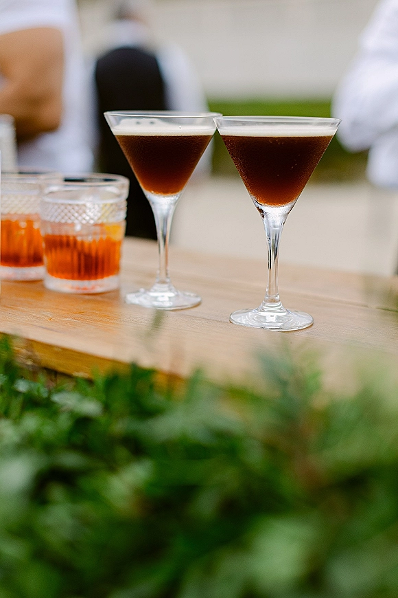 Wedding cocktails in martini and tumbler glasses on a wooden bar top, with greenery and blurred guests against a light wall