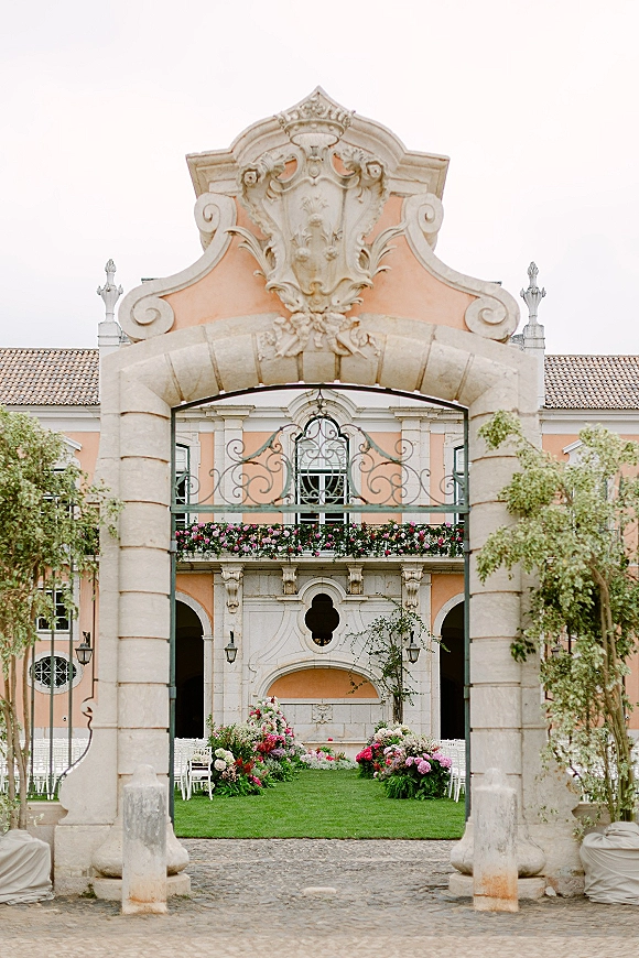 Outdoor ceremony setup with garden ceremony aisle, rows of white chairs, and floral-lined greenery leading to a stone arch and wrought iron gate in a palace courtyard