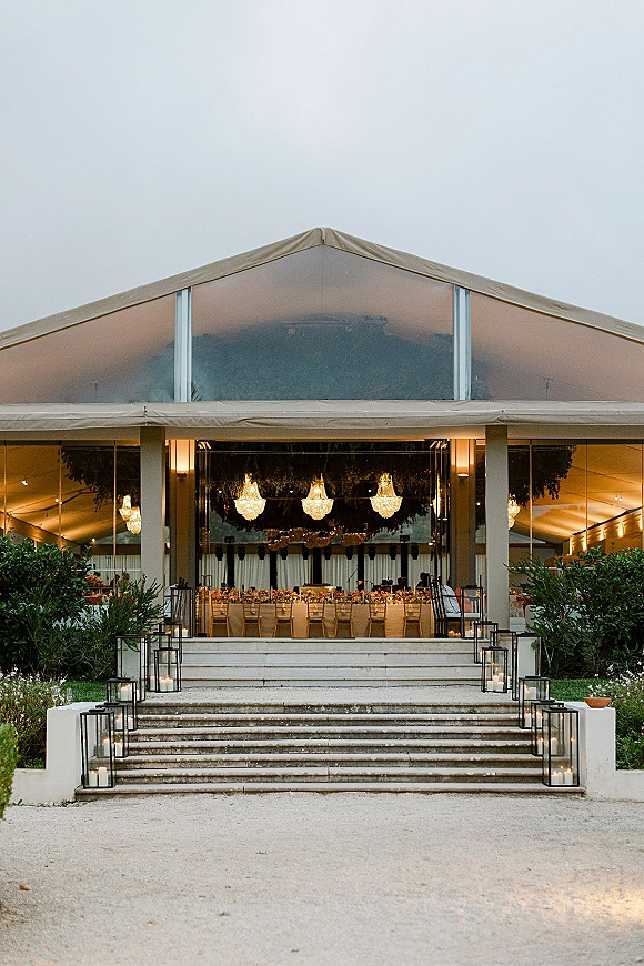 Reception entrance decor with glass lanterns and pillar candles lining steps into a clear-top tent with chandeliers, string lights, and tables beyond