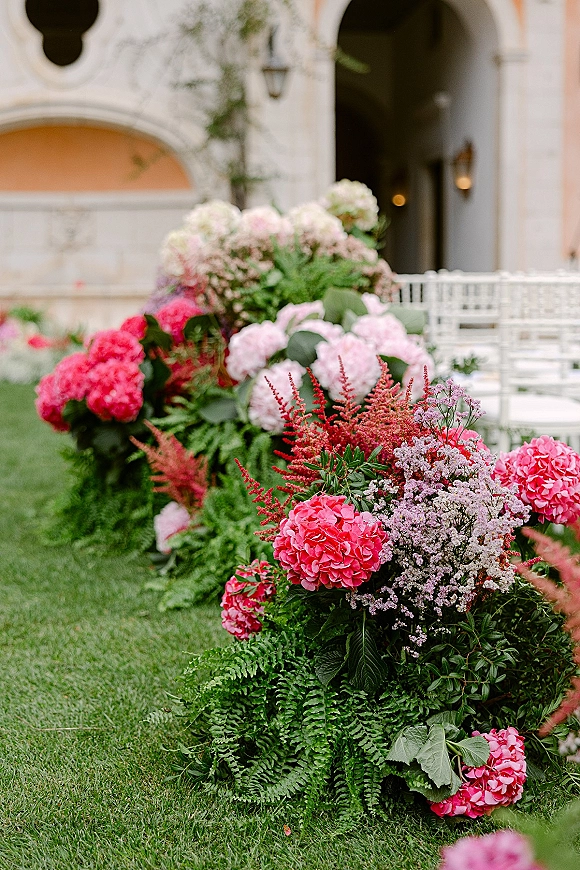 Ceremony aisle flowers with wedding aisle floral arrangements of hydrangeas and ferns lining white chairs on a grass courtyard before an arched facade