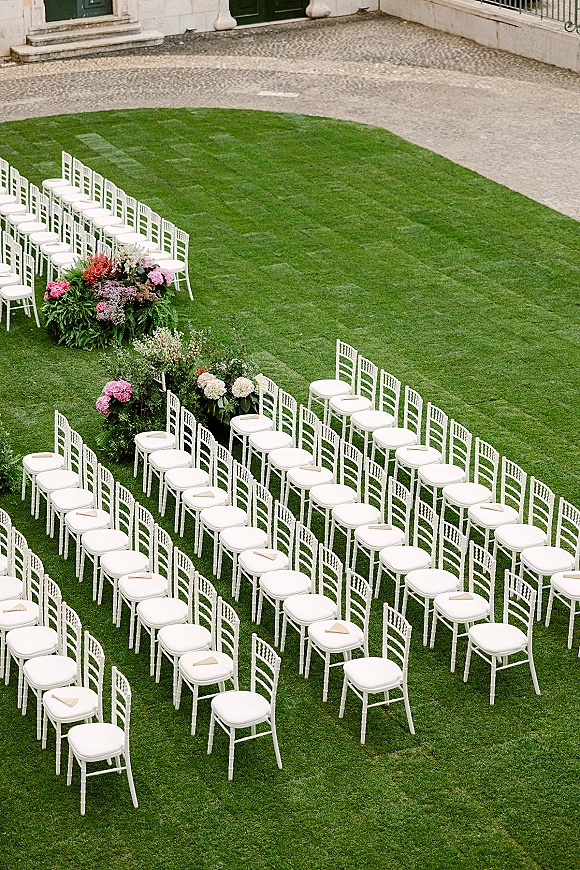 Ceremony seating with white chiavari chairs in a curved outdoor wedding ceremony seating arrangement on a grass lawn with floral aisle markers