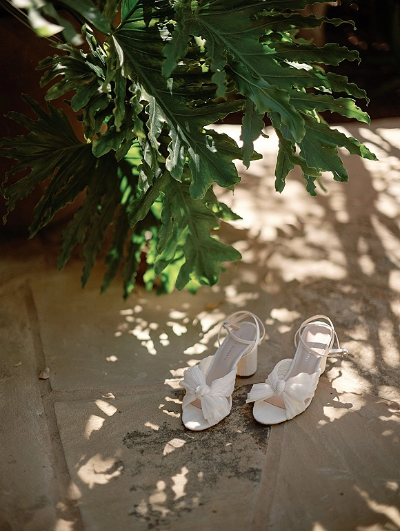 Bridal shoes in white wedding heels with bow accents and ankle straps on a sunlit stone floor with leafy shadows