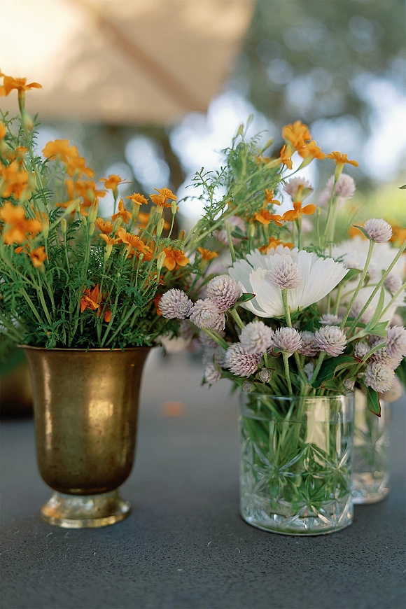Wedding table flowers in wildflower centerpieces with marigolds and white daisies in brass and glass vases under a canopy tent outdoors
