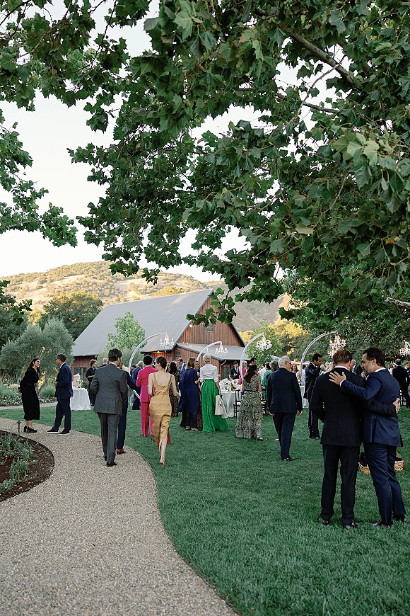 Wedding cocktail hour with outdoor cocktail hour guests mingling around white linen tables under chandeliers near a rustic barn lawn