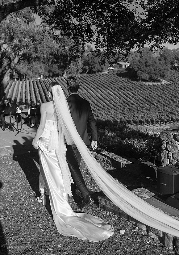Ceremony recessional as bride and groom walk away, her long cathedral veil trailing behind on a vineyard patio with musicians nearby