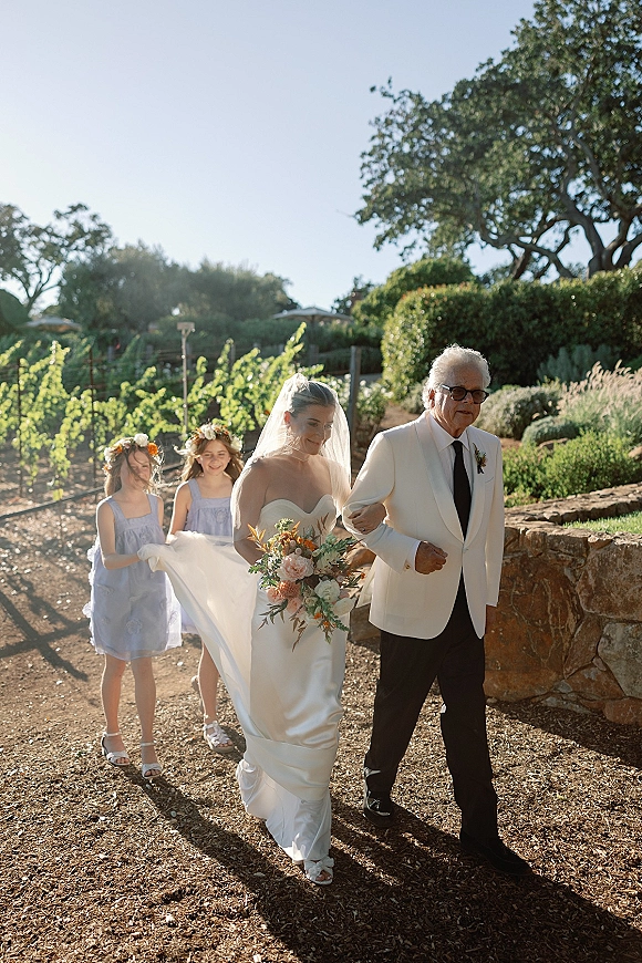 Wedding processional with bride walking down aisle, escorted by her father, bouquet and veil flowing as bridesmaids follow along a vineyard path