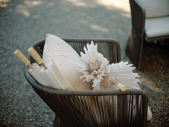 Wedding parasols in a wicker chair beside patio seating, paper parasols stacked as guest shade favors in dappled sunlight on gravel