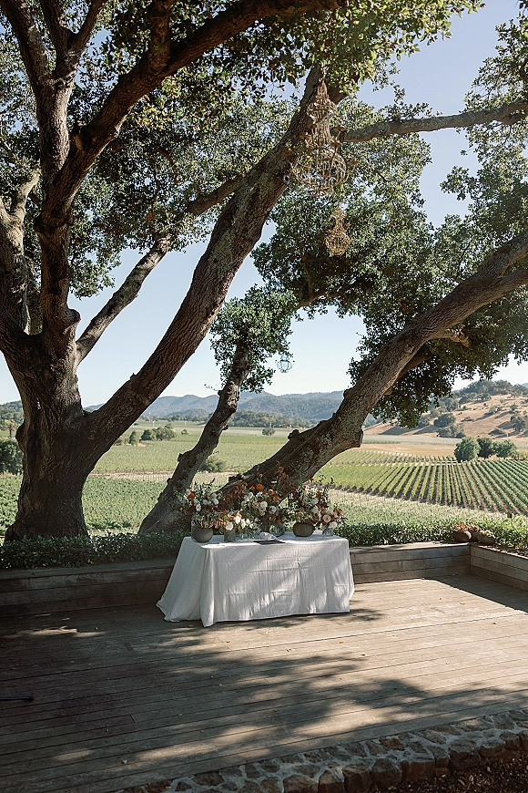 Wedding reception sweetheart table with outdoor sweetheart table florals, white linen and hanging lanterns beneath oak trees overlooking vineyard hills