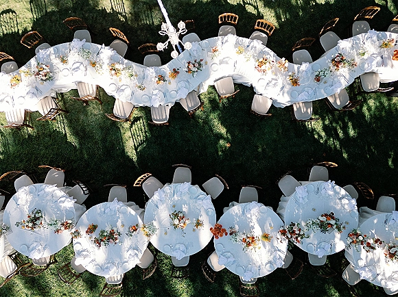 Reception tablescape at an outdoor wedding reception with white linens, floral bud vases, place settings, wooden chairs, and chandelier overhead on lawn