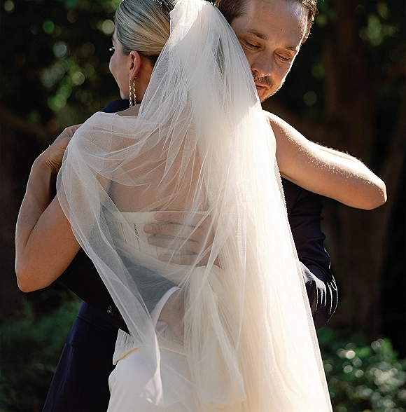 Wedding couple embrace as the groom holds the bride in a strapless dress with a flowing veil in sunlit garden greenery