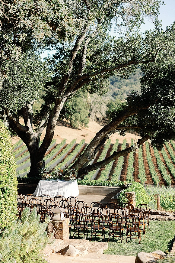Ceremony setup with wood chairs facing a simple altar table and floral arrangement under an oak tree, with chandeliers and vineyard hills beyond